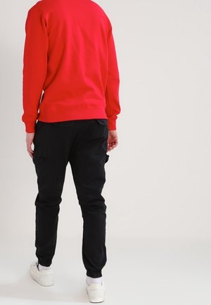 Persoon die een rood shirt met lange mouwen, zwarte cargobroek, witte sokken en witte sneakers draagt, staand tegen een effen lichte achtergrond.