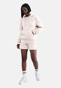 Felpa con cappuccio di colore rosa chiaro con tasca frontale, abbinata a pantaloni corti coordinati. Presenta cuciture bianche e il logo Nike. Indossata con scarpe da ginnastica bianche.