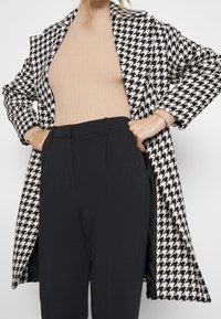 Cappotto a motivo pied de poule in bianco e nero, design lungo, abbinato a un top beige a coste e pantaloni neri a vita alta.
