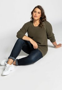 Suéter de punto verde oliva con mangas texturizadas, combinado con jeans de mezclilla oscura y zapatillas blancas. Modelo sentado contra un fondo sencillo.