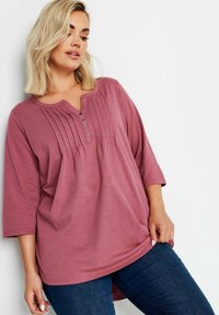 Vaaleanpunainen toppi, jossa on pyöreä kaula-aukko, osittainen nappilista ja pystyröyhelöitä. Pitkät hihat ja rento istuvuus. Yhdistetty tummiin farkkuihin.