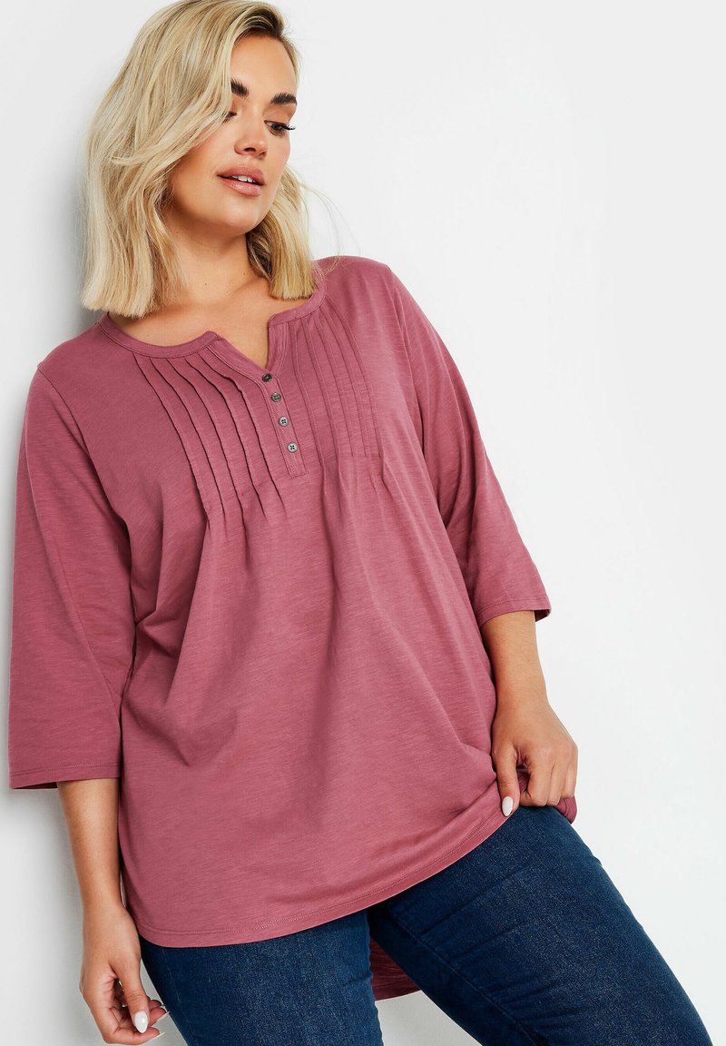 Vaaleanpunainen toppi, jossa on pyöreä kaula-aukko, osittainen nappilista ja pystyröyhelöitä. Pitkät hihat ja rento istuvuus. Yhdistetty tummiin farkkuihin.