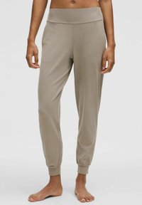 Person, der beiger tapered Jogginghose mit elastischen Bündchen und Bund trägt, barfuß vor einem schlichten hellen Hintergrund steht.