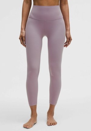 Femme portant des leggings taille haute, de couleur mauve, longueur cheville, debout pieds nus devant un fond uni.