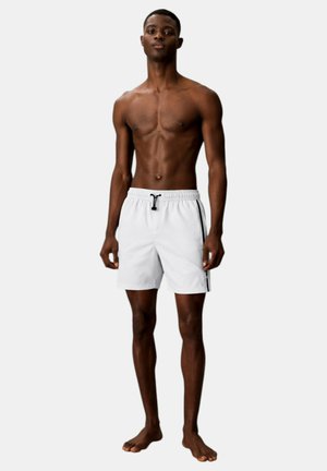 Giovane uomo in piedi a piedi nudi che indossa pantaloncini da bagno bianchi con strisce laterali nere e una cintura regolabile contro uno sfondo semplice.
