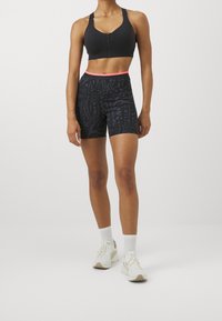 Woman wearing a black zip-front sports bra, black patterned high-waist biker shorts with pink trim, white socks, and white sneakers.