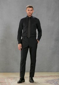 Schwarzes Dressshirt und Krawatte, getragen mit schwarzen, maßgeschneiderten Hosen und polierten Schuhen. Der glatte Stoff und das taillierte Design schaffen eine schlanke Silhouette.