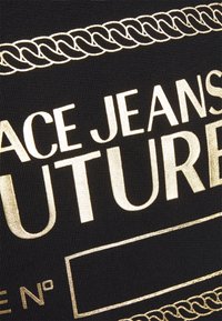 Svart tyg med guldmetallisk text "VERSACE JEANS COUTURE" och dekorativa guldkedjemönster runt topp och botten.