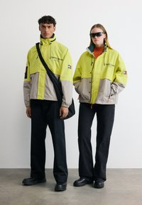 Δύο μοντέλα φορούν κίτρινα-γκρι μπουφάν Tommy Jeans, μαύρο παντελόνι και μαύρα παπούτσια· το ένα κρατά μια μαύρη τσάντα ώμου, το άλλο φοράει μαύρα γυαλιά ηλίου.