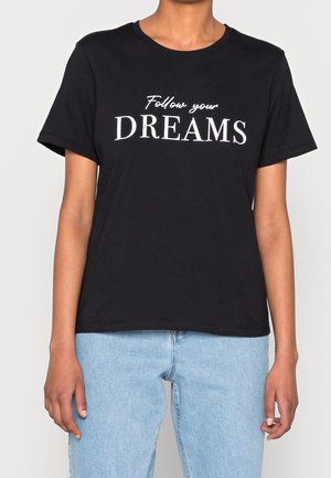 Osoba ubrana w czarną koszulkę z białym napisem "Follow your DREAMS" i jasnoniebieskie dżinsy, stojąca na jednolitym tle.