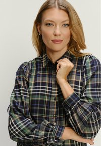Blondínka s modrými očami nosí zelenú, námornícku a béžovú károvanú košeľu s gombíkmi, jednou rukou si drží golier proti jednobarevnému svetlému pozadiu.
