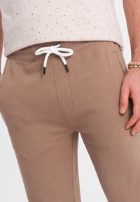 Hellbraune Sweatpants mit einer Kordelzugverschlus, versehen mit schwarzen Kunststoffenden, seitlichen Taschen und einer weichen, strukturierten Oberfläche.