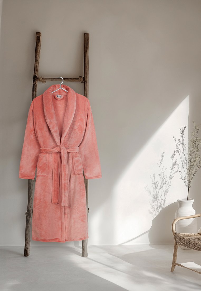 Peignoir de bain en peluche rose corail accroché à une échelle en bois rustique contre un mur beige, avec la lumière du soleil et un vase contenant des branches sèches à proximité.