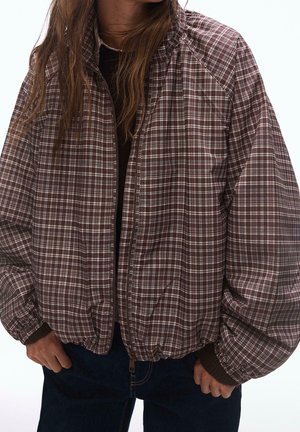 Osoba nosząca brązowo-białą kratkę na kurtce zapinanej na zamek błyskawiczny z elastycznymi mankietami i krawędzią, na ciemnej koszulce, ręce spoczywające blisko kieszeni, na białym tle.