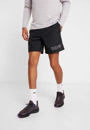 Person trägt schwarze Nike-Shorts, weiße Nike-Socken, dunkle Sneakers und ein hellgraues Langarmshirt und steht vor weißem Hintergrund.