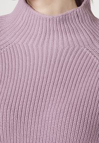 Suéter de punto lila con cuello alto, que presenta una textura de costillas verticales y hombros sin costuras. El material parece suave y grueso.