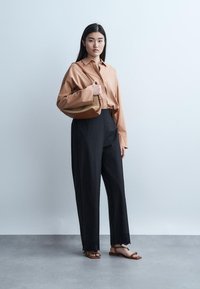 Młoda kobieta stoi na tle gładkiej ściany, ubrana w beżową koszulę na guziki, czarne szerokie spodnie, brązowe sandały, a na ramieniu ma teksturowaną torebkę.