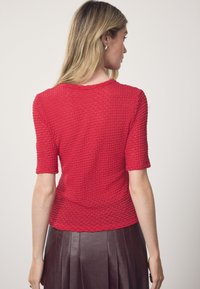 Crveni teksturirani top s kratkim rukavima, okruglim izrezom i skraćenim krojem. Tkanina ima ispupčenu, uzorkovanu površinu, u kombinaciji s smeđom naboranom suknjom.