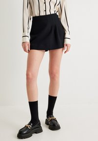 Schwarze, plissierte Shorts mit hoher Taille; kombiniert mit einer gestreiften Bluse. Die Accessoires bestehen aus glänzenden schwarzen Loafers und knielangen Socken.