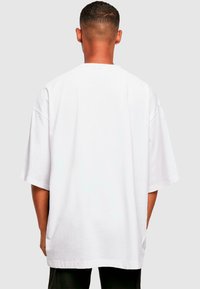 Balta per didelė t-shirt su trumpomis rankovėmis, turinti apvalų iškirpimą ir glotnią tekstūrą. Matomi siūlai palei apatinę dalį ir rankoves.