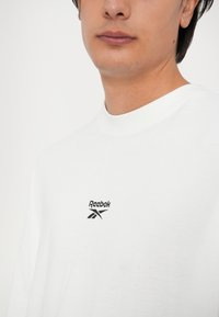 Bijela pamučna majica kratkih rukava s okruglim izrezom. Ima mali crni Reebok logo na prsima, glatka tekstura.
