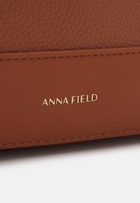 Surface en cuir marron avec des textures lisses et granulées, avec le nom de marque embossé en doré "ANNA FIELD" sur la partie inférieure.