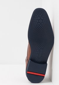 Semelle en cuir brun avec un motif en nid d'abeille, base en caoutchouc bleu marine et accent rouge au talon. Vue latérale sur fond clair.