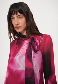 Mulher com cabelo longo e escuro, vestindo uma blusa floral transparente em tons de rosa e preto, com um grande laço amarrado no pescoço, olhando ligeiramente para o lado.