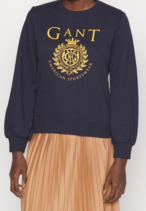 Osoba nosząca granatową bluzę z żółtym logo "GANT American Sportswear" w połączeniu z beżową plisowaną spódnicą, widoczna od szyi do bioder.
