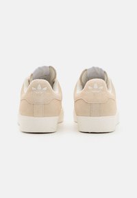 Ténis de camurça bege com solas de borracha branca, apresentando uma marcação discreta no calcanhar e um colar acolchoado para maior conforto.