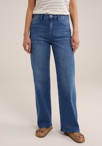 Kvinna i högmidjade blå raka jeans, randig vit och beige topp instoppad, och beige sandaler med öppna tår och spännen.