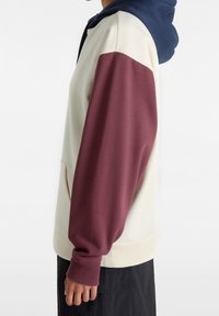 Hooded sweatshirt with a navy hood, cream body, and maroon sleeves. Features ribbed cuffs and a front pocket. Soft fabric texture.