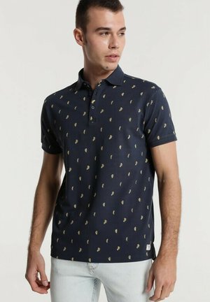 Jeune homme portant un polo bleu marine avec de petits motifs jaunes et un jean clair, regardant sur le côté devant un fond uni.