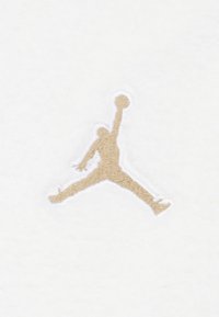 Logo Jumpman brodé beige sur un fond texturé blanc, représentant une figure en plein saut avec un ballon de basket.