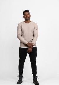 Beige Langarm-Thermooberteil mit strukturiertem Finish, schwarze Akzente an den Ärmeln, kombiniert mit schwarzen engen Hosen und schwarzen Stiefeln.