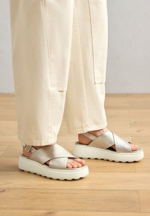 Sandali con incrocio argentato e finitura testurizzata, con una suola spessa bianca e fibbia regolabile alla caviglia, indossati con pantaloni larghi color crema.