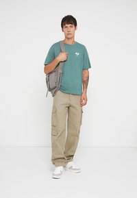 Green cotton t-shirt with a small graphic, paired with beige cargo pants featuring multiple pockets. White sneakers and a gray backpack.
