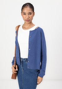 Μπλε πουλόβερ με κουμπιά, μαλακή υφή και μακριά μανίκια, φοριέται πάνω από ένα λευκό ριμπ μπλουζάκι, συνδυασμένο με τζιν φούστα και μια μικρή καφέ τσάντα.