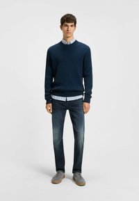 Navy ribbed sweater over a light blue striped shirt, paired with dark denim jeans and gray sneakers with a gum sole. Simple, casual design.