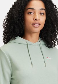 Sudadera de color verde menta con cordón y extremos de color plateado. Presenta un pequeño logo bordado en el pecho. Textura de tela suave y lisa.