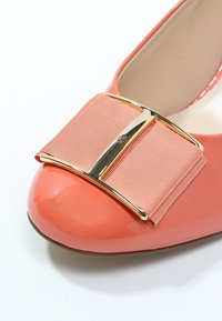 Chaussure plate en cuir verni corail avec un large nœud pastel rose et un accent en métal doré au bout. Texture lisse, forme arrondie.