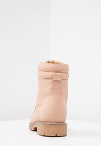 Beige ankelboot med vadderad krage, texturerat blommönster på ovandelen, tjock sula med mönster, sedd bakifrån på en vit yta.