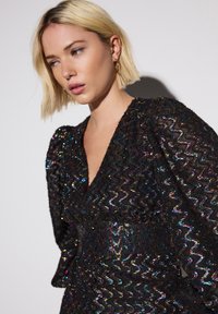 Zwarte jurk met glinsterende stof met golfpatroon. Heeft een diepe V-halslijn en volumineuze mouwen. Subtiele iriserende accenten door de hele jurk.