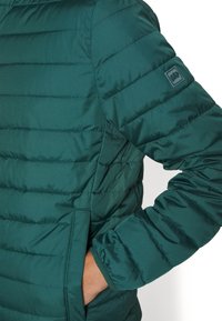 Teal puffer jacket with horizontal stitching, featuring a side pocket and elasticized cuffs. Small logo patch on the sleeve.