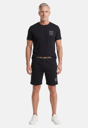 Un uomo con capelli grigi indossa una maglietta nera CR7 e pantaloncini con scarpe da ginnastica bianche e sta in piedi, rivolto in avanti, su uno sfondo bianco.