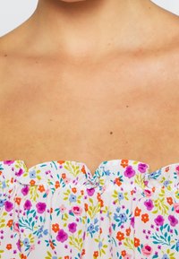 Vit blommig topp med färgglada tryck i rosa, lila och orange. Har insamlad halsringning med smala volanger och mjuk textur.