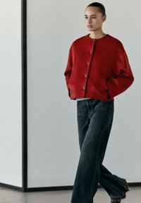 Cardigan oversize rosso con bottoni, realizzato in lana, abbinato a jeans scuri a vita alta in denim. Tessuto liscio, scollo rotondo e vestibilità dritta.