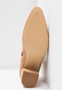 Chaussure en daim beige à bout pointu et talon bloc vue de la semelle, taille 37 indiquée, reposant debout contre des surfaces blanches.