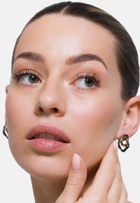 Orecchini astratti oro e nero indossati da un modello con una pelle liscia, che mostra un trucco naturale e una mano che tocca l'orecchino.