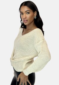 Cremiger, gestrickter Pullover mit V-Ausschnitt und gedrehtem Frontdetail, langärmlig und mit lässiger Passform. Glatte Textur, leichtes Material.